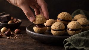 Een hand plakt baci di dama met pure chocolade op een donker bord, met hele hazelnoten en een blok chocolade ernaast
