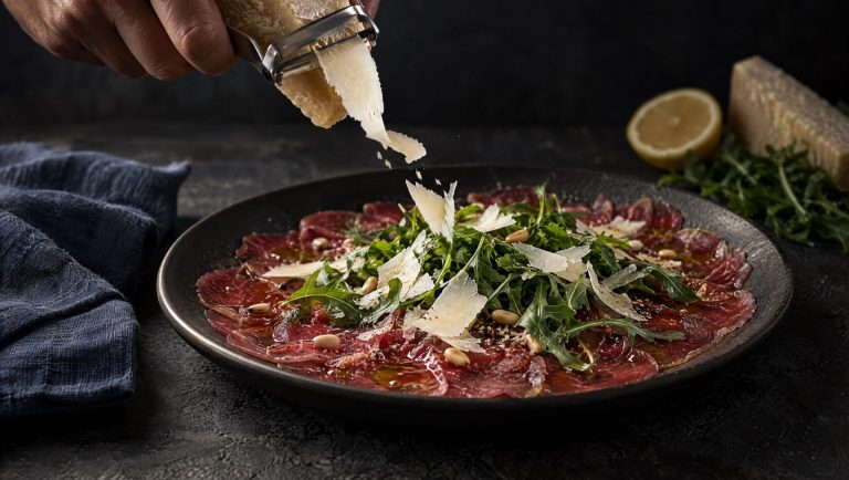 Carpaccio van rund met rucola, pijnboompitten en Parmigiano die erover geschaafd wordt, met citroen en kaas ernaast