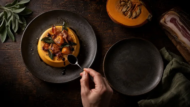 Hartige panna cotta van pompoen met pancetta en balsamico op een donkere tafel in Emilia, met rauwe pompoen en salie
