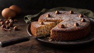 Aangesneden hazelnoottaart met poedersuiker en hele hazelnoten op een donker bord, met rauwe hazelnoten en eieren
