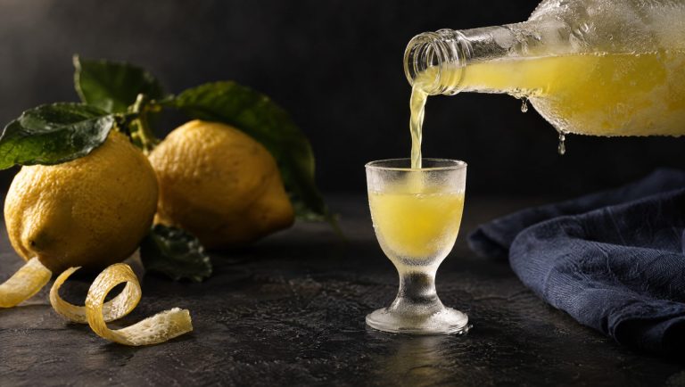 IJskoude limoncello wordt uit een beslagen fles in een klein glaasje geschonken, naast citroenen met blad en schillen