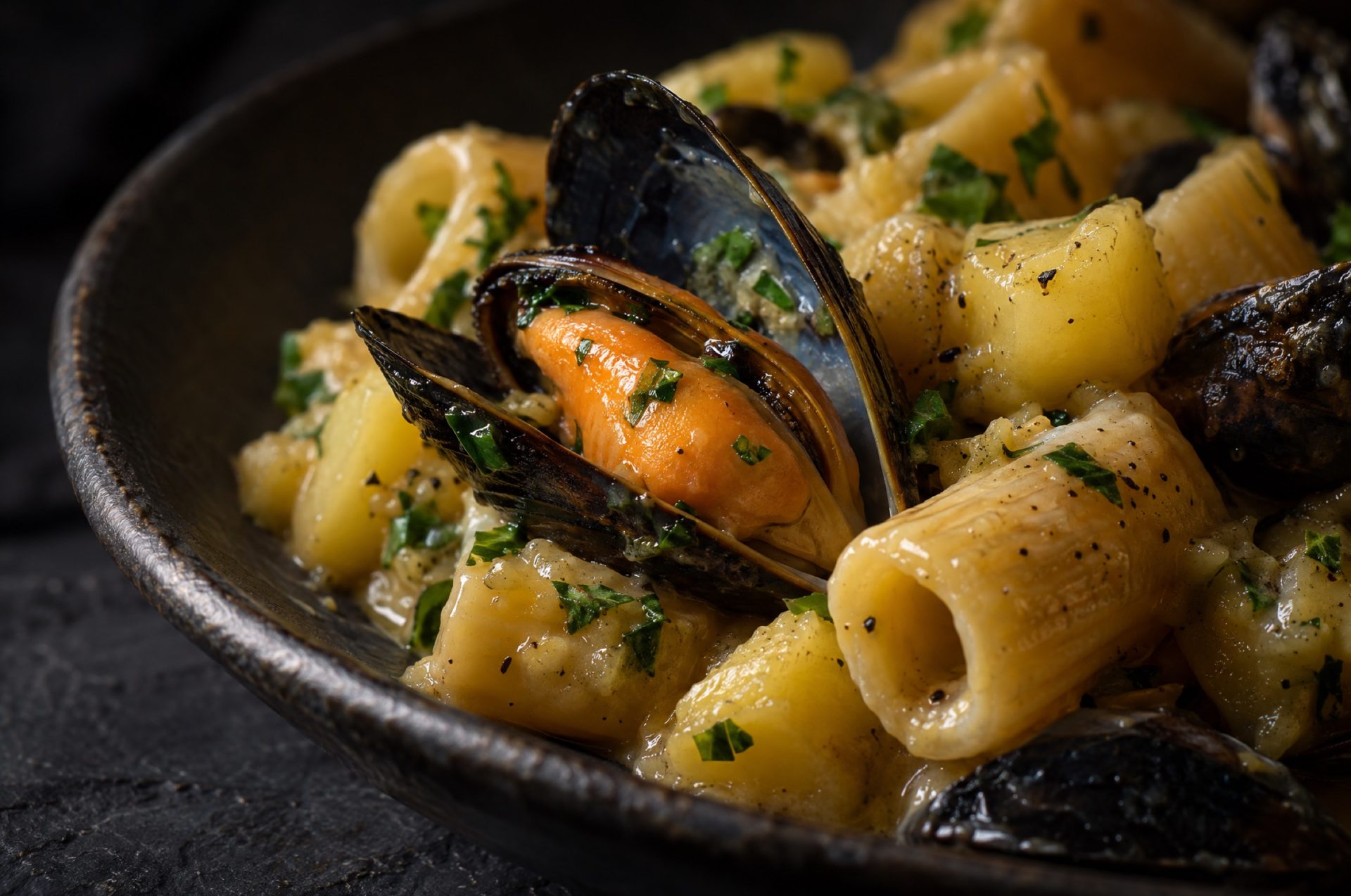Detail van pasta met aardappel en mosselen: glanzende saus over mezze maniche, een open mossel en gehakte peterselie