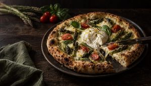 Pizza met asperges en burrata op een donker bord, met groene asperges, kerstomaatjes en basilicum ernaast