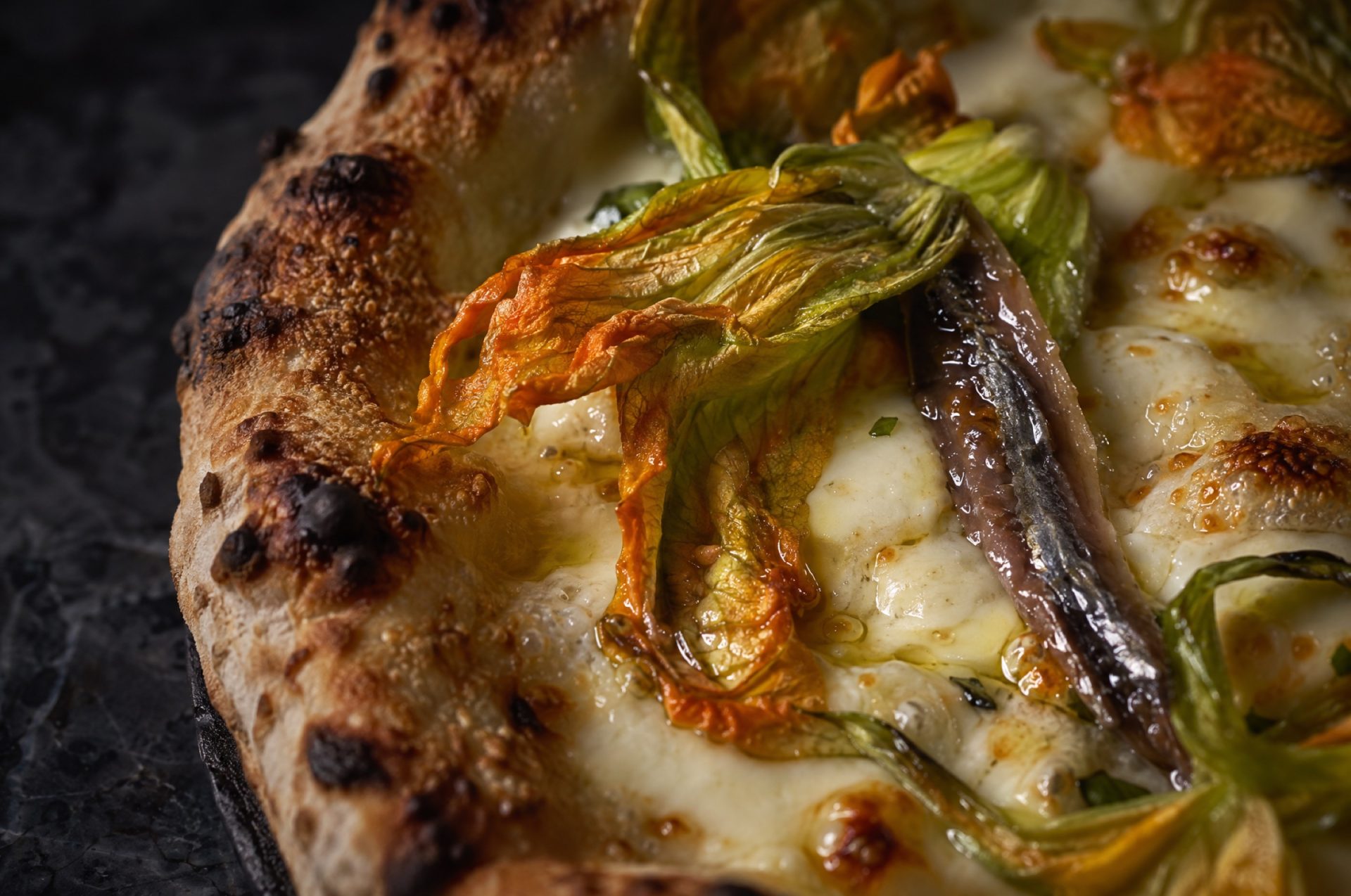Close-up van pizza met courgettebloemen: geslonken oranje bloemen, ansjovis en gesmolten mozzarella op een goudbruine bodem