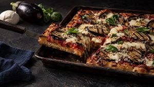 Pizza parmigiana op een bakplaat op een donkere tafel, met een aubergine, mozzarella en basilicum ernaast
