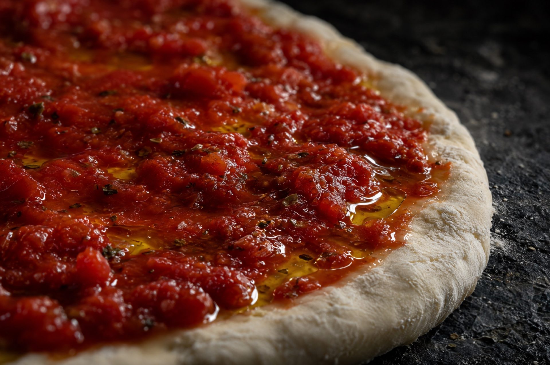 Pizzasaus maken zonder pan: close-up van rauwe saus op een pizzabodem, met stukjes tomaat en glanzende olijfolie