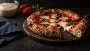 Aangesneden pizza met poolish op een houten plank, met gepelde tomaten, basilicum en een kom voordeeg