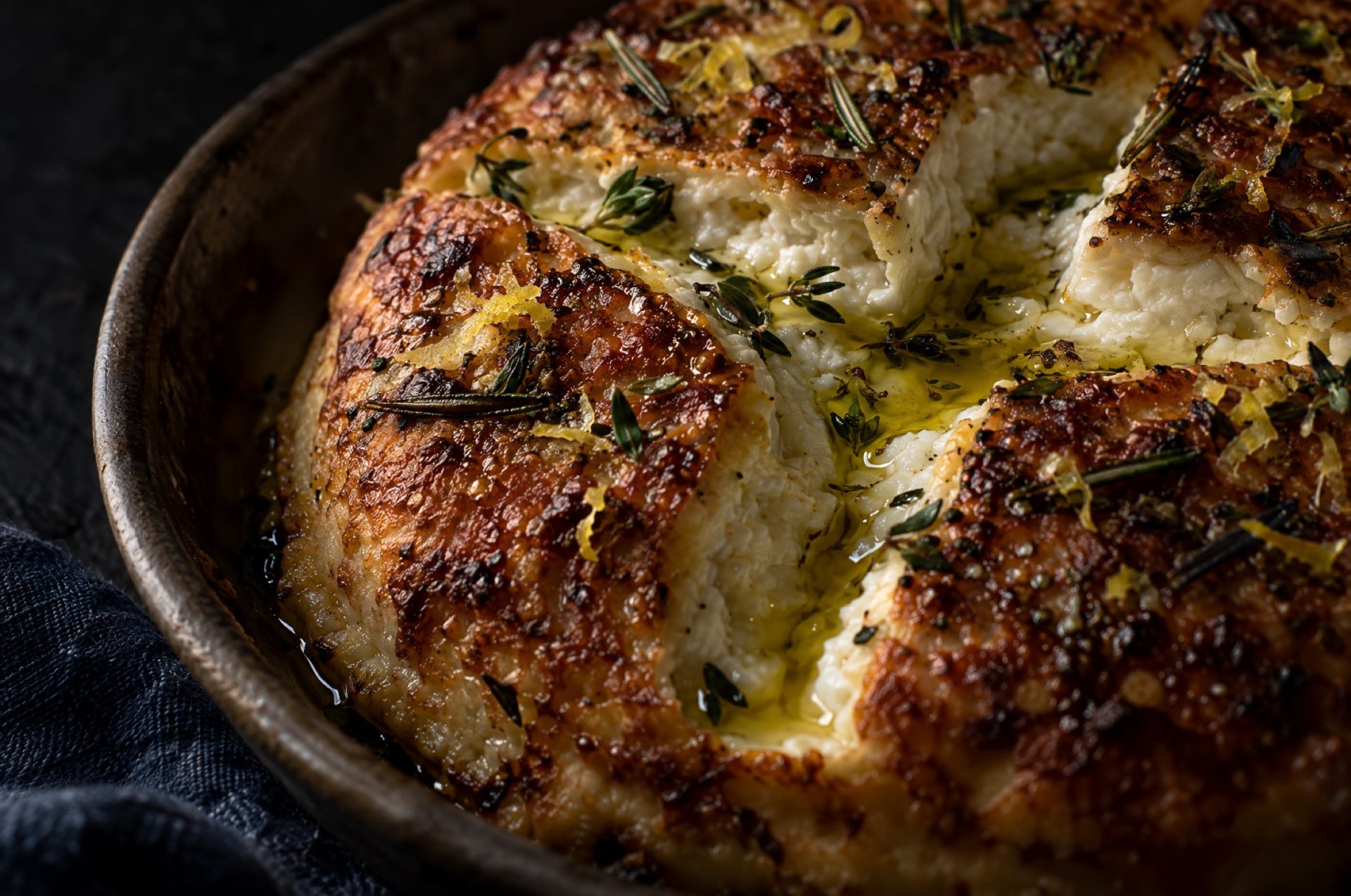 Detail van ricotta uit de oven: de goudbruine korst met kruisinsnede, glanzende olijfolie en stukjes tijm