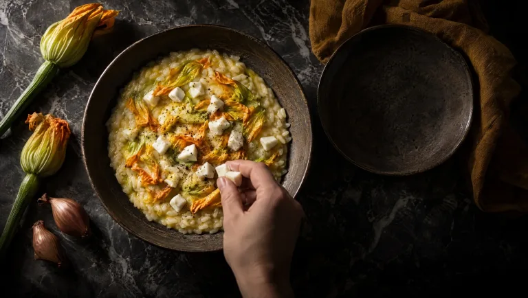 Risotto met courgettebloemen en mozzarella in een diep bord op een donkere tafel, met verse bloemen en sjalotten