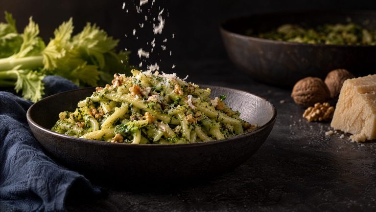 Selderijpesto met trofie in een donkere kom, met vallende Parmigiano, bleekselderij, walnoten en een stuk Parmigiano